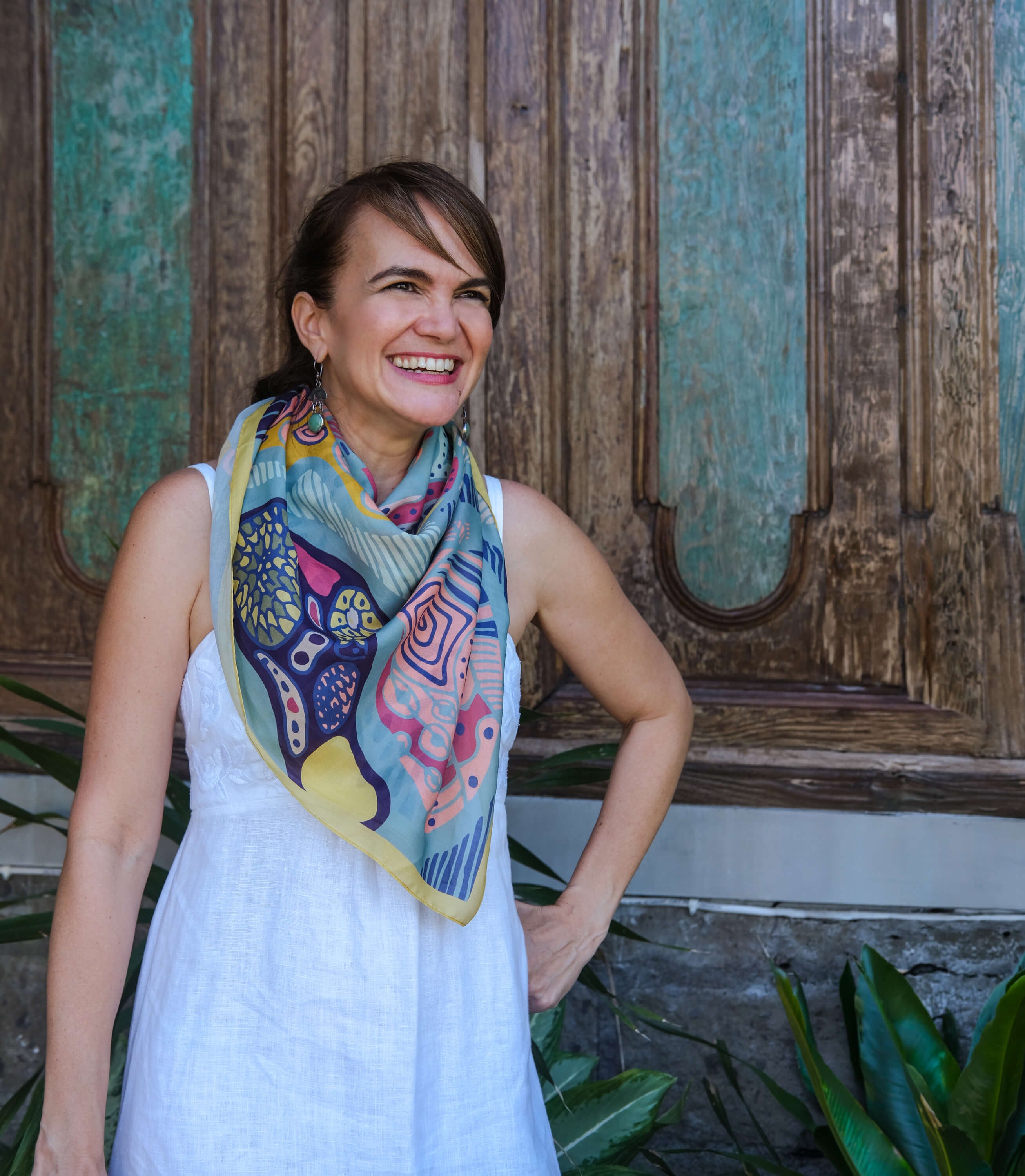 A woman wearing a white sleeveless dress and a colorful patterned scarf, standing in front of a rustic wooden door with green plants at the bottom.